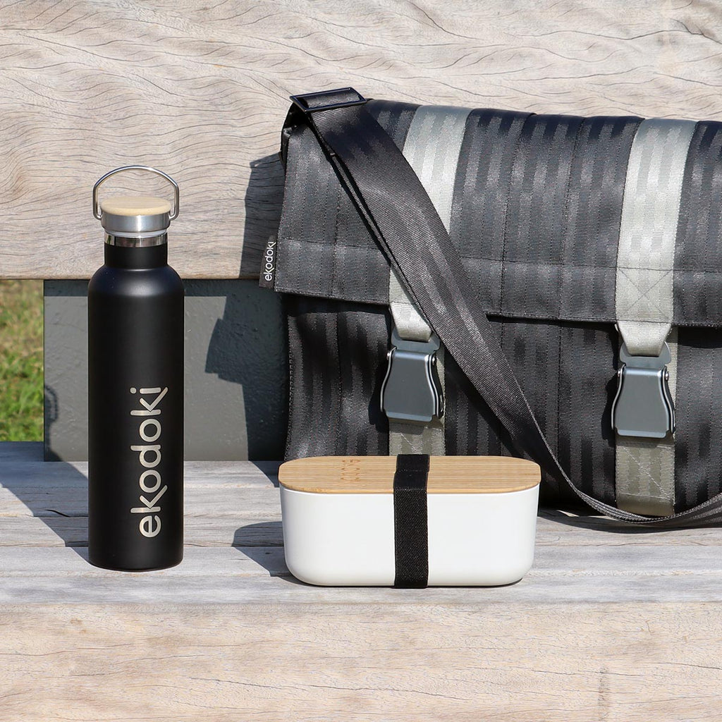 black reusable bottle, white bamboo lunchbox with black strap, and black seatbelt messenger bag, all from the brand eKodoKi