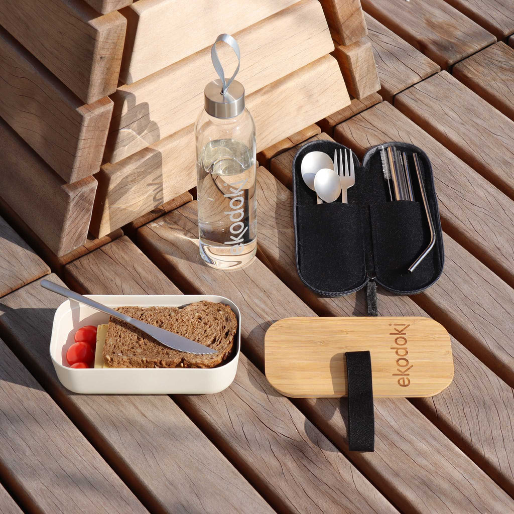 3 eKodoki accessories for lunch on-the-go: a 700ml white lunch box (with tomatoes and a sandwich in it) and its bamboe lid; a reusable glass bottle (filled with water), and a stainless steel cutlery set and its grey/black real wool felt travel pouch.